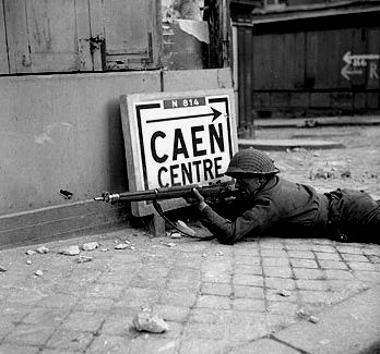 photograph of a soldier laying with a gun on the ground behind a building